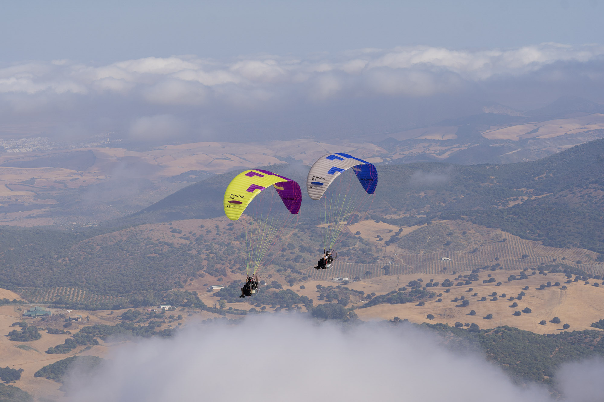 Two paramotor pilots flying together above the clouds over the Andalusian countryside